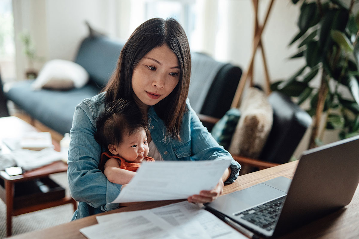 woman researching on computer and looking at document while holding baby