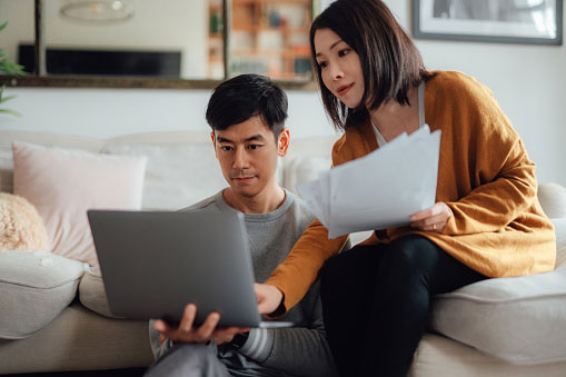 Man sits on floor next to coffee table holding a laptop. Woman on the right is sitting on the couch holding papers and peering on to laptop with man.