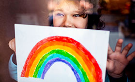 young child holding their drawing of a rainbow up to the window