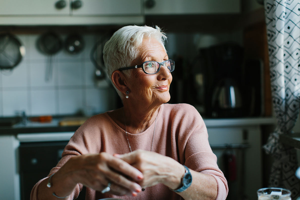 Older woman sitting at her dining table in her kitchen, smiling while looking out the window on the right.
